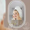 A woman with a towel on her head is looking into a lighted mirror, touching her face. Produktbild-Vorschau