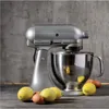 A silver KitchenAid stand mixer with a metal bowl sits on a counter, with lemons and eggs in front of it. Produktbild-Vorschau