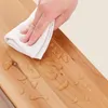A hand is wiping a wooden surface with a white cloth, showing water droplets on the surface. Produktbild-Vorschau