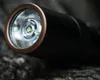 Close-up of the head of a black flashlight with a copper ring around the lens, resting on a textured surface. Náhled obrázku produktu