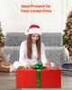 A woman in a Santa hat is wrapping a large red gift box with a green ribbon, sitting on the floor in front of a grey couch. Christmas trees are visible in the background. Anteprima dell’immagine prodotto
