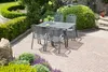 A garden dining set with a table and four chairs on a paved patio, surrounded by lush greenery and landscaping. Produktbild-Vorschau