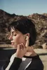 A woman in profile is adjusting an earbud in her ear, with a desert landscape in the background. Anteprima dell’immagine prodotto