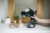 A hand holding a black garment steamer over a white dresser, with decorative items in the background. Voir les images du produit