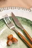 A close-up shot of a fork and knife with wooden handles resting on a green plate, with hazelnuts in the foreground. Produktbild-Vorschau