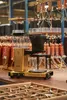 A Moccamaster coffee maker is shown in a factory setting, with many copper coils in the background. Náhľad obrázku produktu