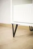 A close-up shot of a black hairpin leg attached to a white piece of furniture, resting on a light brown wooden floor. Podgląd zdjęcia produktu