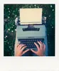 A person's hands typing on a vintage typewriter outdoors, with grass and small white flowers in the background. Voir les images du produit