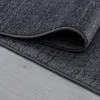 A close-up view of a dark gray rug with a textured pattern, folded over to show the underside and edge. The rug is placed on a light gray wooden floor. Voir les images du produit