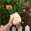 A hand holding a plush toy of tulips in a pot, resting on a wooden fence with a brick wall and greenery in the background. Produktbild-Vorschau