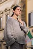 A woman wearing a grey knitted sweater stands outdoors in front of a building. Produktbild-Vorschau