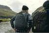 Two people wearing backpacks walk through a rocky, natural landscape near a stream. Náhled obrázku produktu