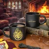 Two black Hogwarts mugs are displayed on a wooden table in front of a fireplace, with books and glasses nearby. Náhled obrázku produktu