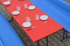 A red table set with white plates, glasses, and cutlery, covered by blue tarps on the sides. Produktbild-Vorschau
