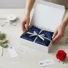 Hands are shown closing a white gift box containing a navy blue item wrapped with silver ribbon. Other gift-wrapping items like ribbon, tags, and a red rose are also visible. Produktbild-Vorschau