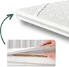 A close-up view of a mattress topper, showing its layers and texture, with a hand lifting a section to reveal the internal structure. Produktbild-Vorschau