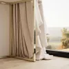 A close-up view of a beige curtain hanging from a gold-colored frame, with a blurred outdoor landscape visible through a window. Náhľad obrázku produktu