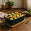 A casserole dish filled with vegetables and sauce is placed on a wooden table, with a fork and a bottle of olive oil in the background. Produktbild-Vorschau