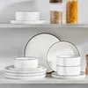 A set of white ceramic dishes with black trim is displayed on a shelf. Voir les images du produit