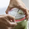 Hands are shown holding a glass jar with a red-banded lid, and a metal tool is inserted through the lid into the contents. Podgląd zdjęcia produktu
