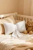 A white bow-shaped pillow and a teddy bear are placed on a bed with beige bedding. Produktbild-Vorschau