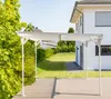 A white aluminum carport is shown in front of a modern white house with a green lawn and a large green bush. Podgląd zdjęcia produktu