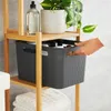 A hand is reaching for a grey storage basket on a wooden shelf. The basket contains several bottles. There is also a plant and a bottle of liquid on the shelf above. Stacked white towels are on the shelf below. Produktbild-Vorschau