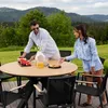A couple enjoying a meal outdoors at a round wooden table with chairs, set against a scenic mountain backdrop. Produktbild-Vorschau
