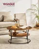A round coffee table with two tiers is placed in a living room setting, next to a sofa. The table has a rustic wood finish and black metal legs. It is decorated with a pitcher of water, a glass, a book, and a small tray with snacks. Produktbild-Vorschau
