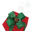 A close-up of a red Christmas gift box with a large green glitter bow, a pine cone, and red berries. Podgląd zdjęcia produktu