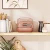 A pink hand mixer with its accessories stored inside a clear plastic case, placed on a wooden shelf next to books and decorative vases. Voir les images du produit