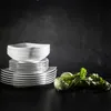 A stack of white square plates and bowls sits on a black surface in front of a black, textured background. Two kohlrabi vegetables with green leaves are placed to the right of the plates. Náhľad obrázku produktu