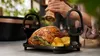 A person is seasoning a roasted chicken with potatoes and rosemary in a kitchen setting. Produktbild-Vorschau