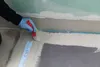 A person wearing blue gloves is applying a sealant to a wall and floor joint with a brush. Produktbild-Vorschau