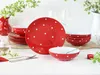 A set of red and white polka dot tableware is arranged on a white surface, with some greenery and flowers in the background. Náhľad obrázku produktu