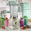 A vintage-style blender is shown on a kitchen counter with several colorful smoothies and a bowl of ice in front of it. A retro refrigerator is visible in the background. Produktbild-Vorschau
