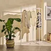 A white metal clothing rack with several garments hanging on it, placed next to a large potted plant in a woven basket. The scene is set in a room with textured walls and soft lighting, casting shadows. Produktbild-Vorschau