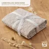 A folded beige blanket with a white ribbon tied around it, placed on a wooden floor with dried flowers. Anteprima dell’immagine prodotto
