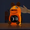 A child's alarm clock shaped like a mouse is sitting on a wooden surface in a dimly lit room. A hand is gently touching the top of the mouse's head. Produktbild-Vorschau