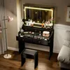 A vanity table with a lighted mirror, drawers, and makeup products, along with a stool and a floor lamp in a room. Produktbild-Vorschau