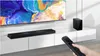 A soundbar and subwoofer are placed on a white media console in front of a television displaying a colorful abstract image. A hand is holding a remote control, pointing it towards the soundbar. Náhled obrázku produktu