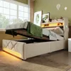 A bed with its storage compartment open, revealing space underneath. The bed has a tufted headboard and is illuminated by soft yellow LED lights. A green blanket is draped over the open mattress. Produktbild-Vorschau