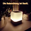 A bedside table with a glowing cube-shaped lamp, a plant, and a mug. Produktbild-Vorschau