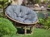 A round cushion on a rattan chair in a garden setting with a pond in the background. Produktbild-Vorschau