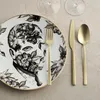 A close-up of a plate with a black and white floral design and a man's face, along with gold cutlery and wine glasses in the background. Produktbild-Vorschau