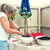 A woman is shown tending to a small tree in a pot on a table outdoors. Produktbild-Vorschau