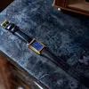 A rectangular gold-cased watch with a blue face and black leather strap is laid diagonally on a dark, textured surface. Produktbild-Vorschau