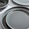 A close-up view of stacked gray ceramic plates and a bowl, with a white tablecloth underneath. Produktbild-Vorschau