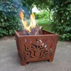 A square metal fire pit with a flame pattern cut into its sides, containing burning logs and flames, sits on a paved patio surrounded by greenery. Podgląd zdjęcia produktu