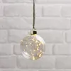 A clear, well-lit product shot of a decorative glass ornament filled with fairy lights, suspended against a white brick wall. Produktbild-Vorschau